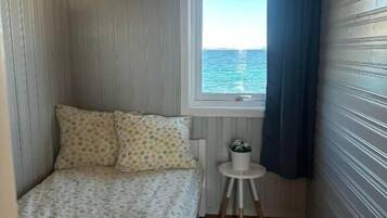 A small, narrow room with a bed, a window with a view of the sea, and a small table with a plant.
