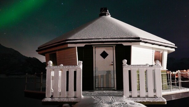 A snow-covered building with a white fence and a door, set against a night sky with visible stars and the aurora borealis. - Fjordcamp AS (Straumsjøen)