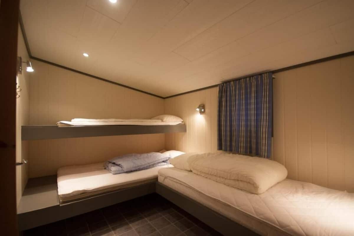 A small, compact room with bunk beds and a single bed. There is a window with curtains, a wall-mounted light, and a door.