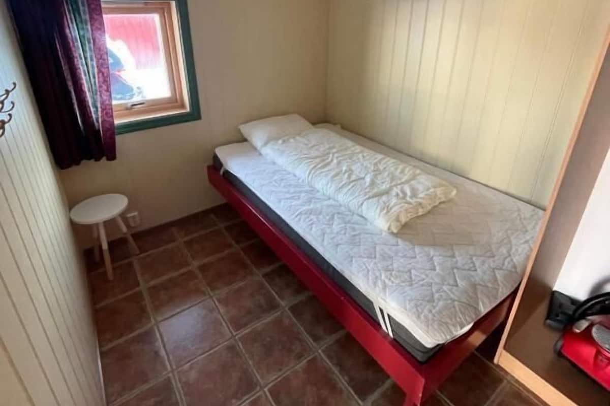 A small, single-bed room with a red frame, a small window with a curtain, and a tiled floor.