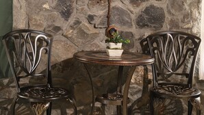 A stone wall, two ornate metal chairs, a small round table with a potted plant, and a hanging light fixture. - The Ranch (Goris)