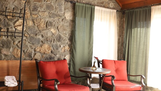 A room with stone walls, wooden ceiling beams, and two red upholstered chairs around a small table. - The Ranch (Goris)