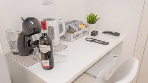 A white desk with a coffee maker, a bottle of wine, a coffee bar, a remote control, and a potted plant. - Pampas Roma (Rome)
