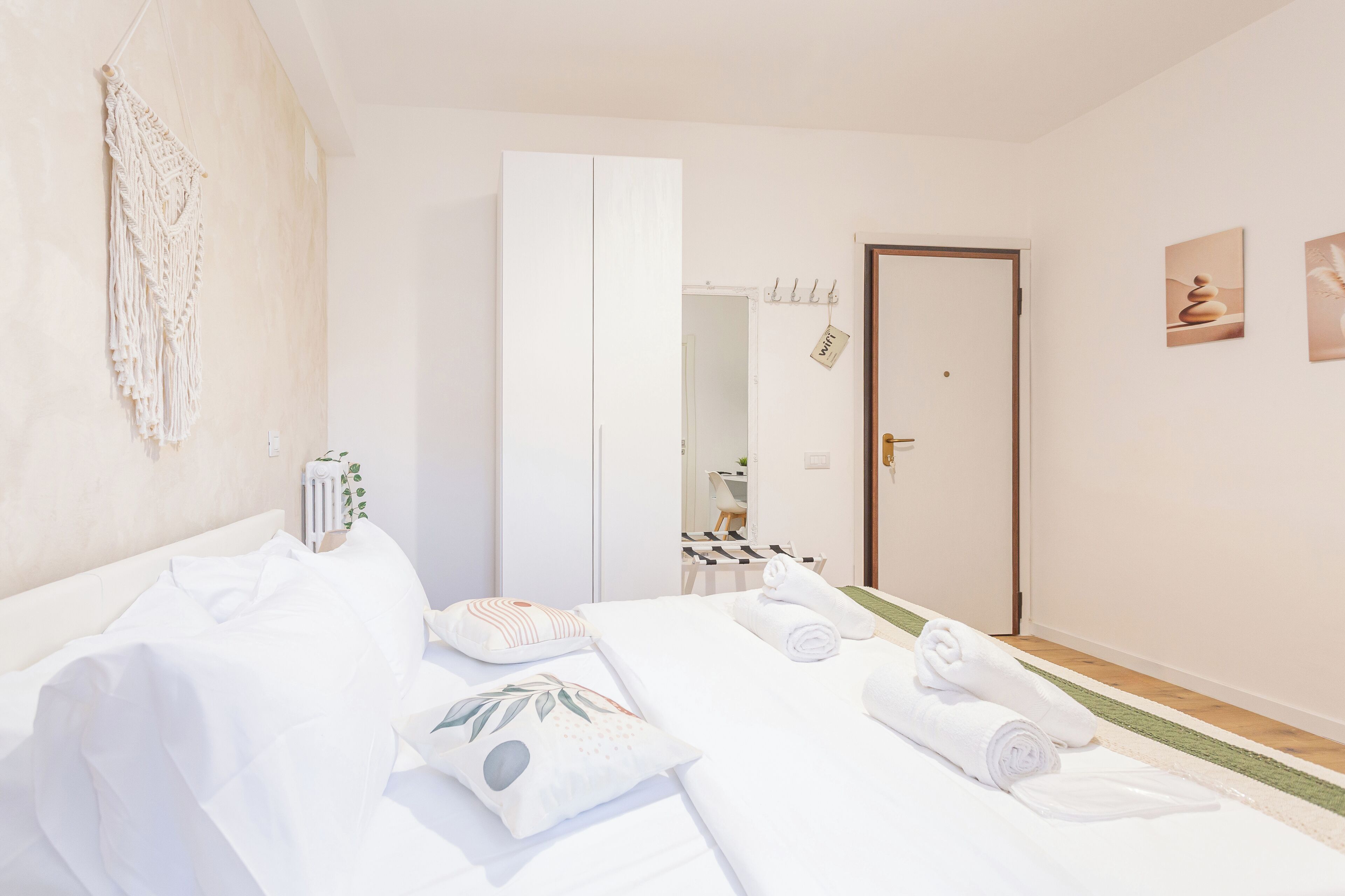 Ein Hotelzimmer mit Bett, weißen Leinentextilien, einer Wandweberei aus Makramee, einem Spiegel und einer Tür mit Handtuchhalter.