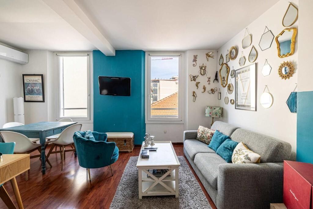 A modern living room with a blue accent wall, a flat-screen TV, a dining area with a wooden table and chairs, and a grey sofa with decorative pillows.