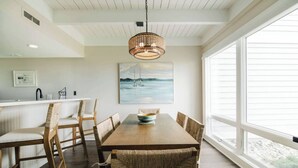 Een eetruimte met een houten tafel, rieten stoelen, een hanglamp en een groot raam met uitzicht op zee.