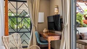 Une pièce avec un bureau en bois, une télévision, des fauteuils en osier et une vue sur la verdure par une fenêtre ornée d’un motif géométrique.