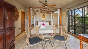 Une chambre à coucher avec un lit à baldaquin, deux fauteuils en osier, une petite table et un ventilateur de plafond.