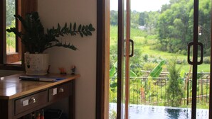 Een kamer met een houten bureau, een plant in pot en uitzicht op een weelderige tuin door een schuifdeur.