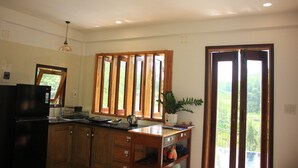 A kitchen with wooden cabinets, a black refrigerator, a ceiling fan, a window with blinds, and a view of greenery outside. - Nomad Hill Hue - Spacious View Countryside (Thị xã Hương Thủy)