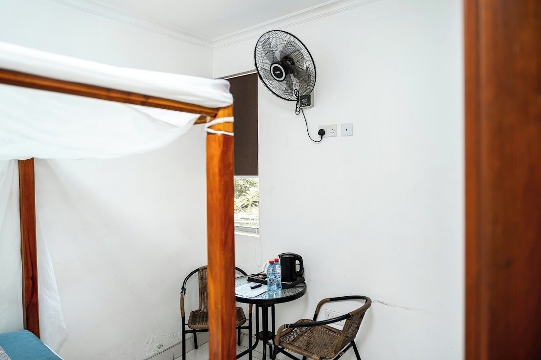 Une petite pièce avec un lit, une table et des chaises, un ventilateur et une fenêtre.