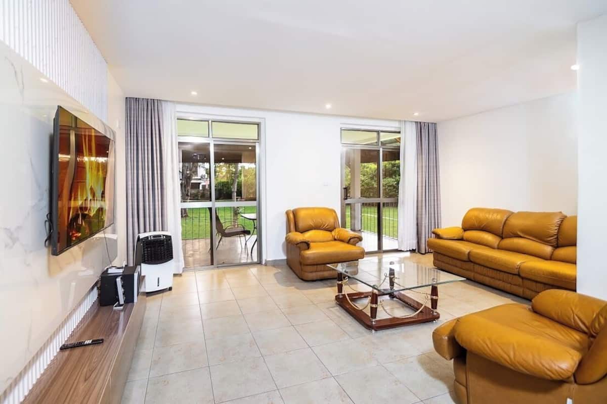 A modern living room with a large sofa, a coffee table, and a flat-screen TV mounted on the wall. There are two windows with curtains, and a glass door leading to an outdoor area.