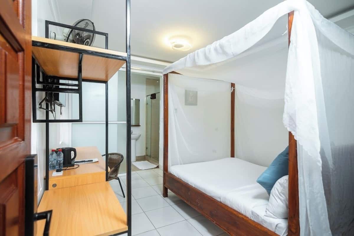 A single bedroom with a wooden bed, a desk with a chair, and a shelf with a fan.