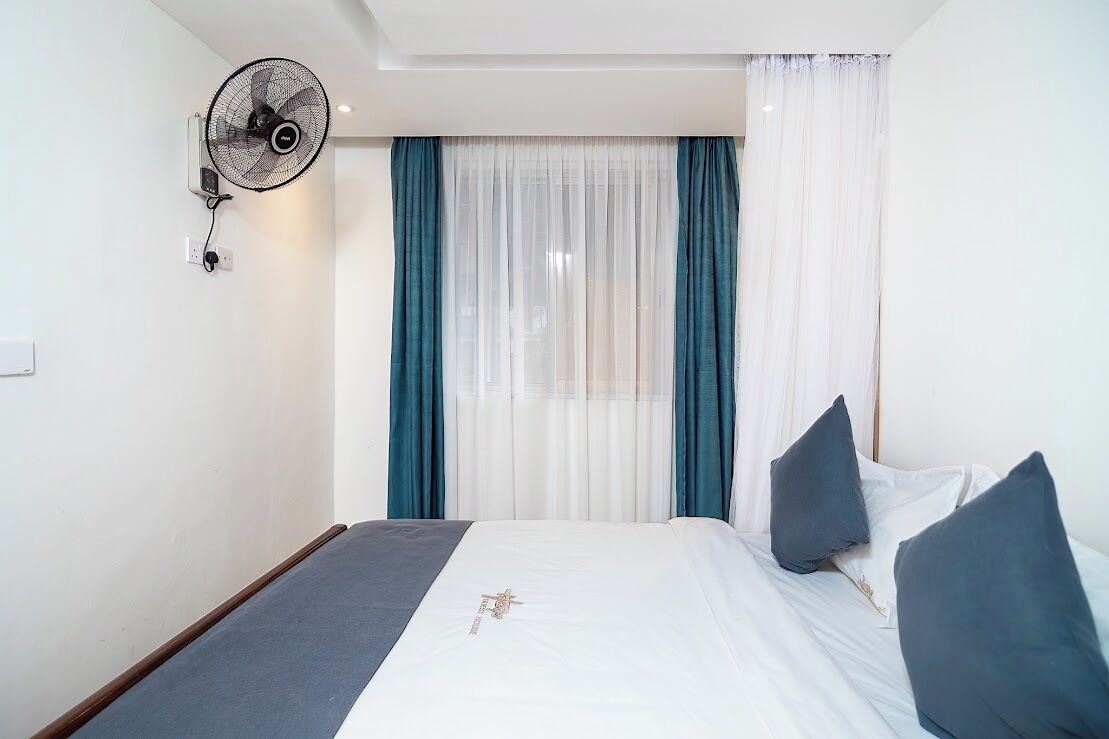 A hotel room with a bed, a wall-mounted fan, and sheer curtains.
