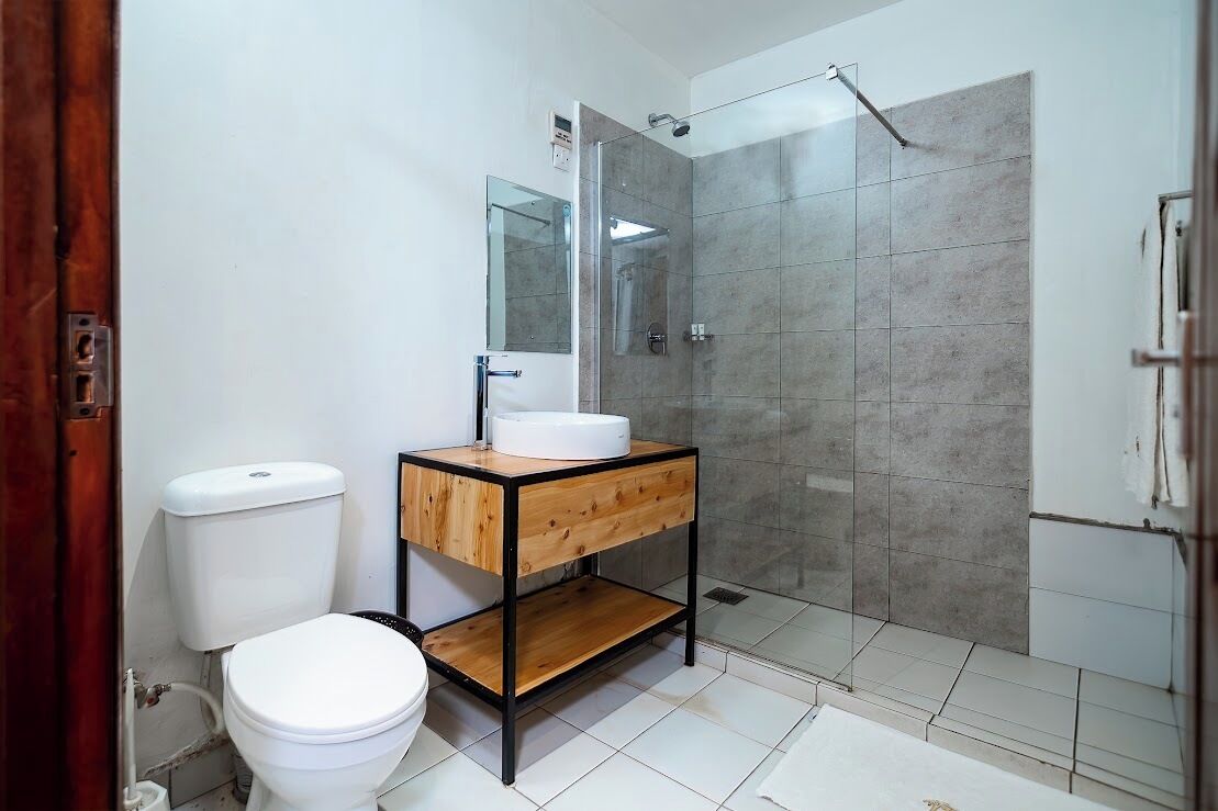 Une salle de bain comprenant des toilettes, un meuble-lavabo en bois avec un évier, un miroir et une douche séparée par une paroi en verre.
