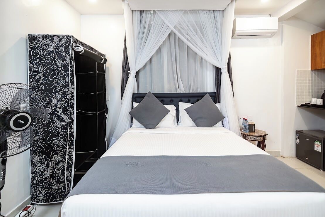 Un dormitorio con cama, ventilador, armario y una mesita con una botella.