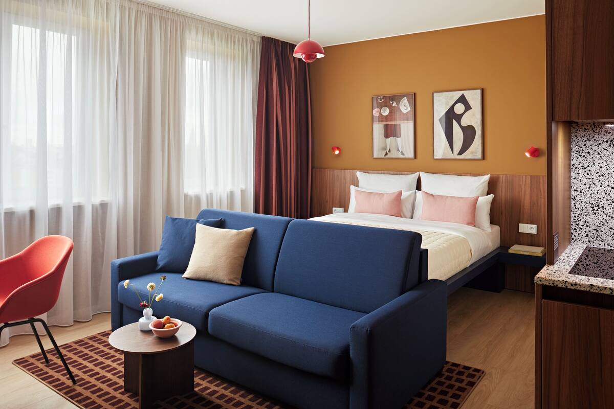 A hotel room with a blue sofa, a red chair, a bed with white and pink bedding, and artwork on the wall.