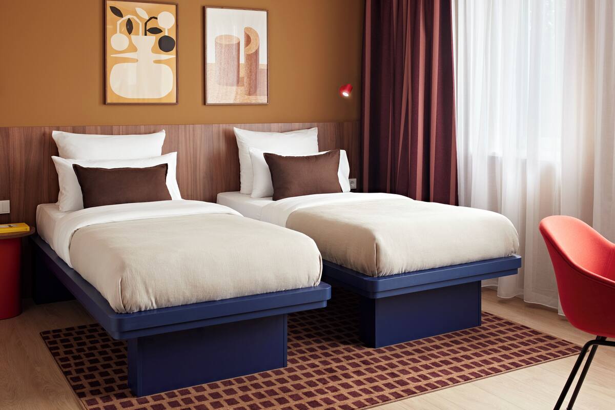 A hotel room with two beds, a red chair, and artwork on the wall.