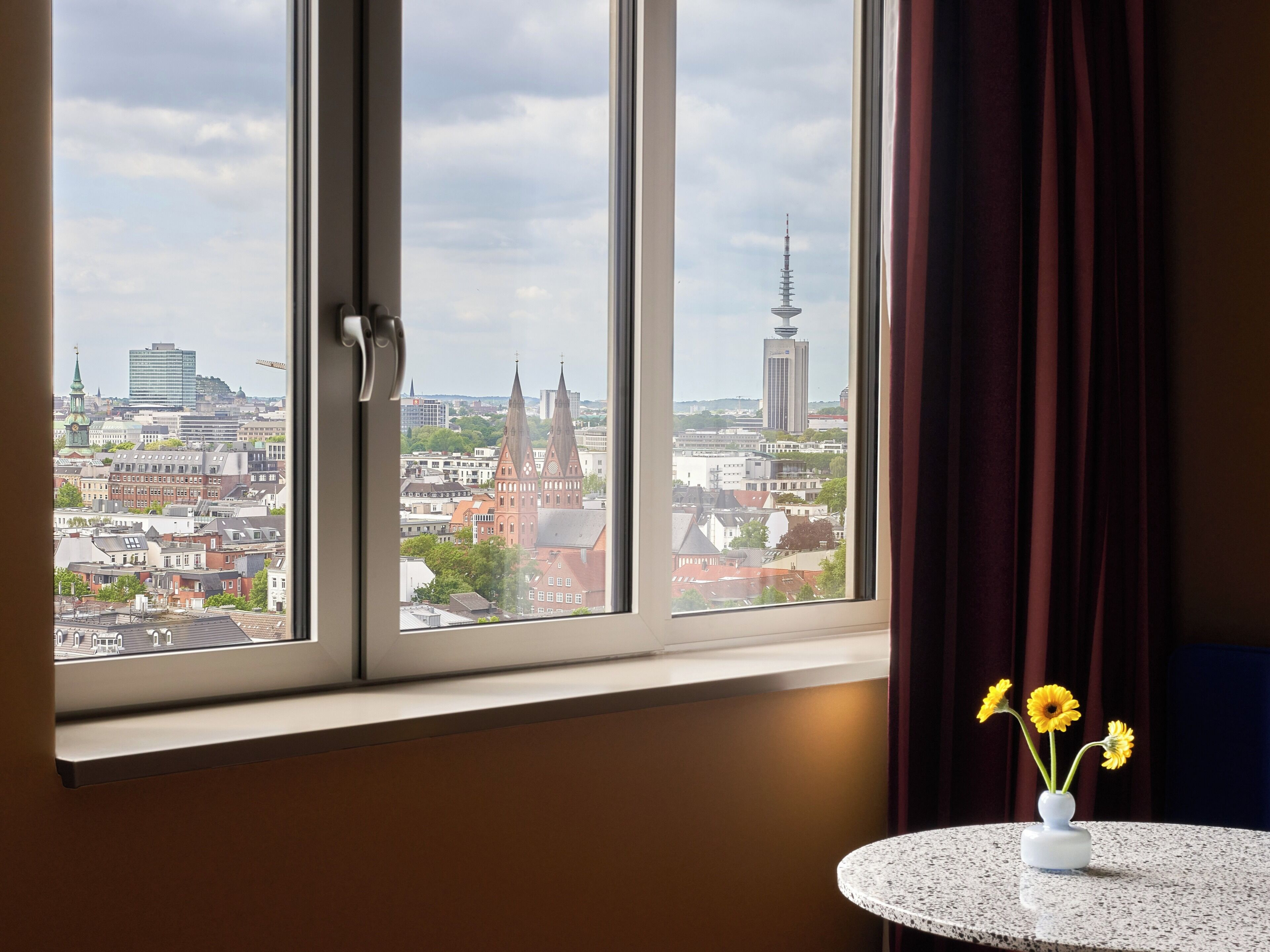 A room with a window overlooking a cityscape, a round table with a vase of flowers, and a curtain partially drawn.