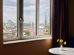 Una habitación con ventana que da a un paisaje urbano, una mesa redonda con un jarrón de flores y una cortina entreabierta.