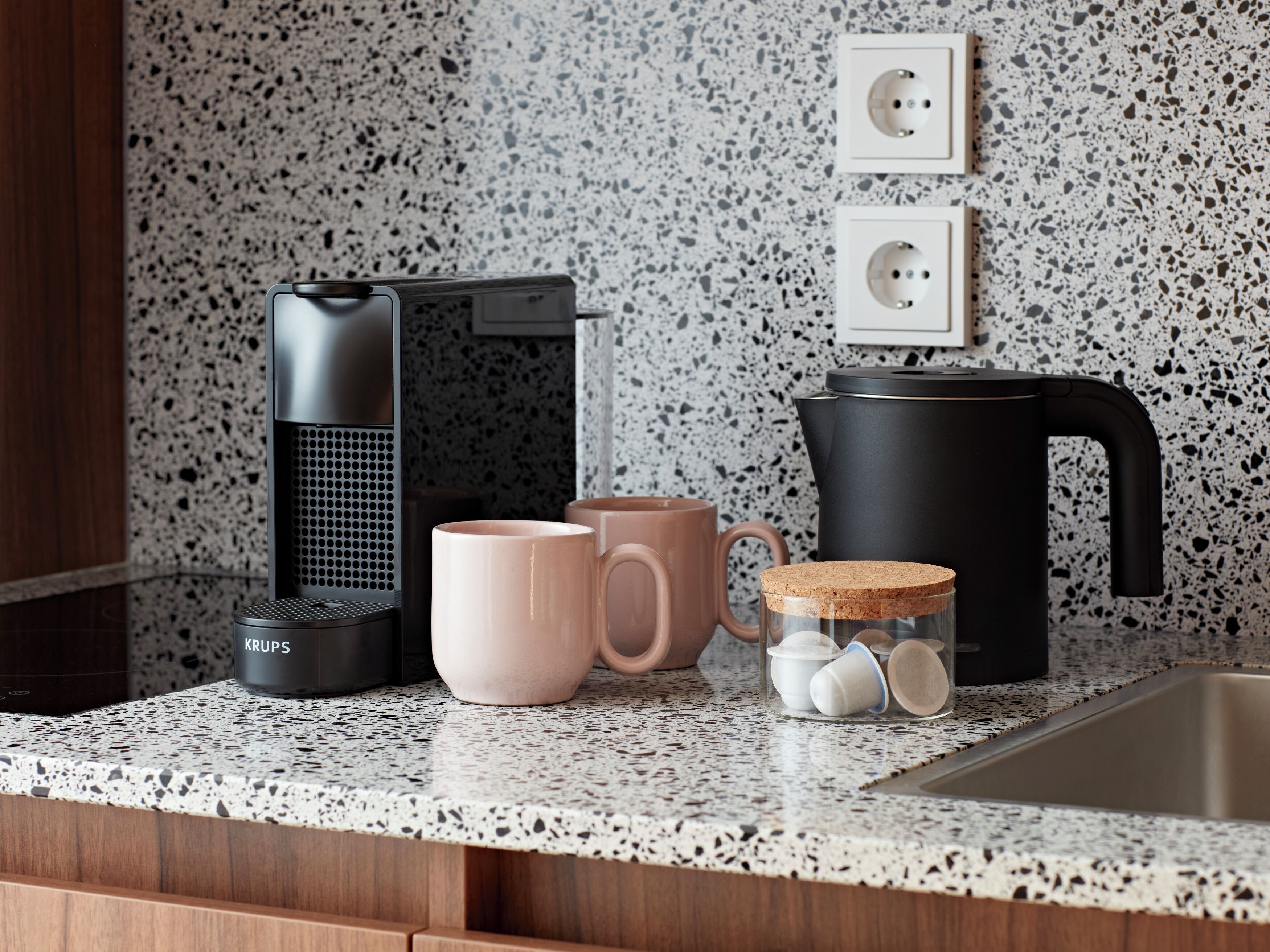 Una cocina con cafetera, dos tazas, un tarro de bolsitas de té y un hervidor sobre una encimera con motas.