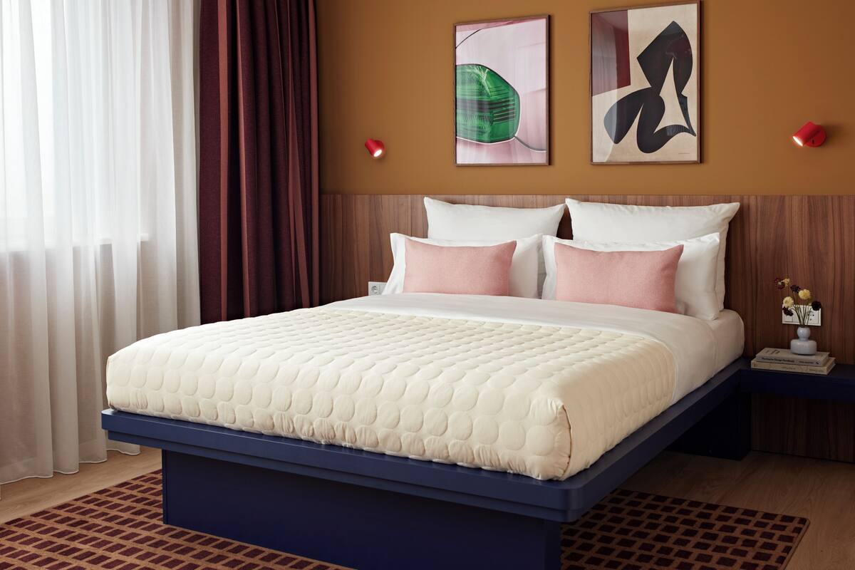 A neatly made bed with a blue base and white bedding, a nightstand with a vase of flowers, and two abstract paintings on the wall.