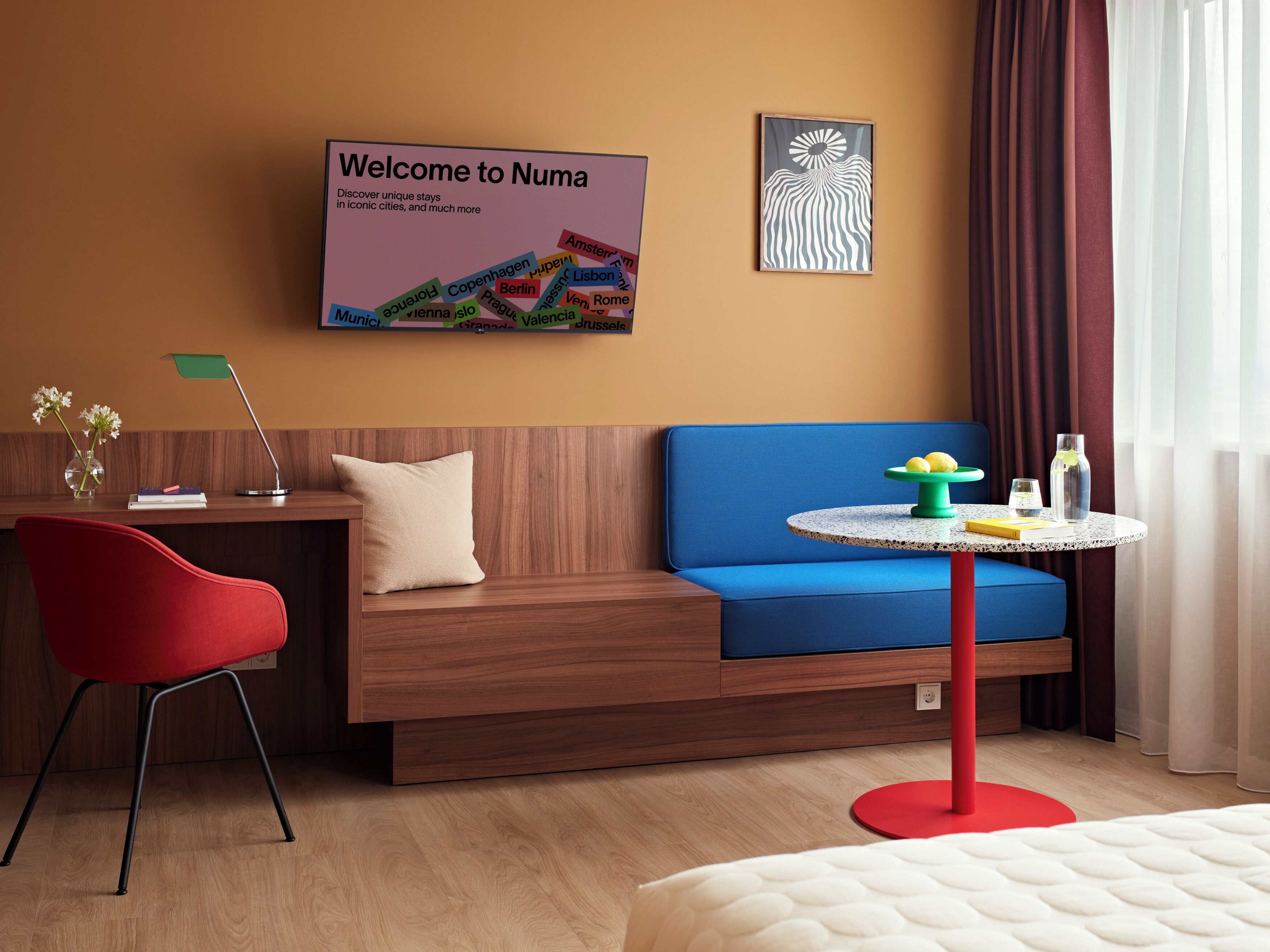 Ett modernt hotellrum med en soffa i trä, en röd fåtölj, ett litet runt bord med en grön fot och en säng med en vit madrass.