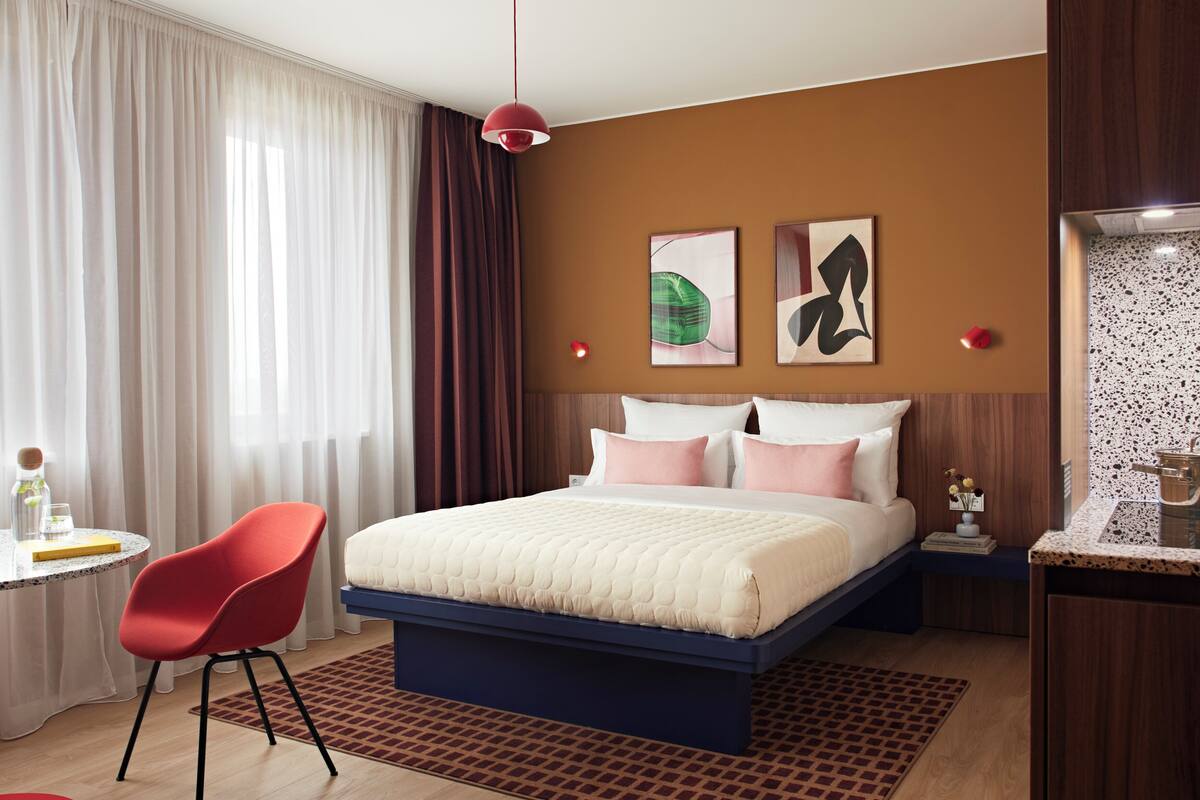 A hotel room with a bed, a red chair, a small table with a glass and a book, and a nightstand with a lamp.