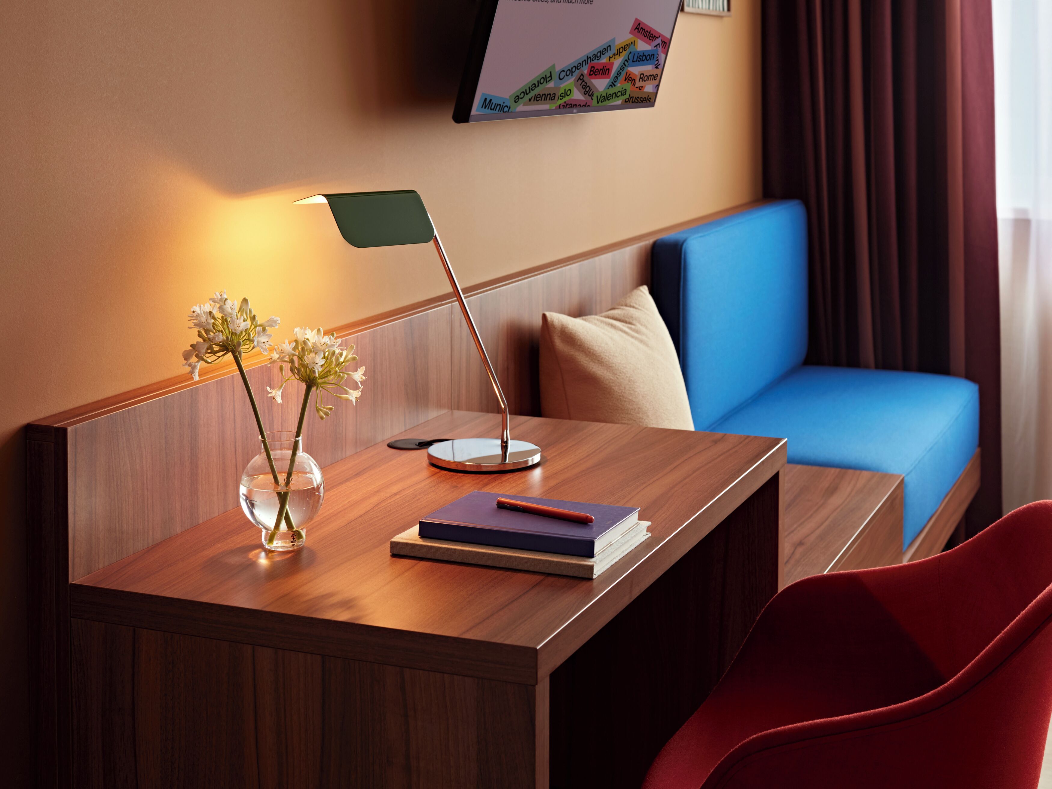 A hotel room with a wooden desk, a vase with flowers, a lamp, a blue sofa, and a TV mounted on the wall.