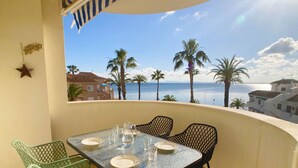 A balcony with a table set for two, overlooking a coastal view with palm trees and buildings. - Beautiful apartment fully refurbished with lovely sea views. (La Manga del Mar Menor)