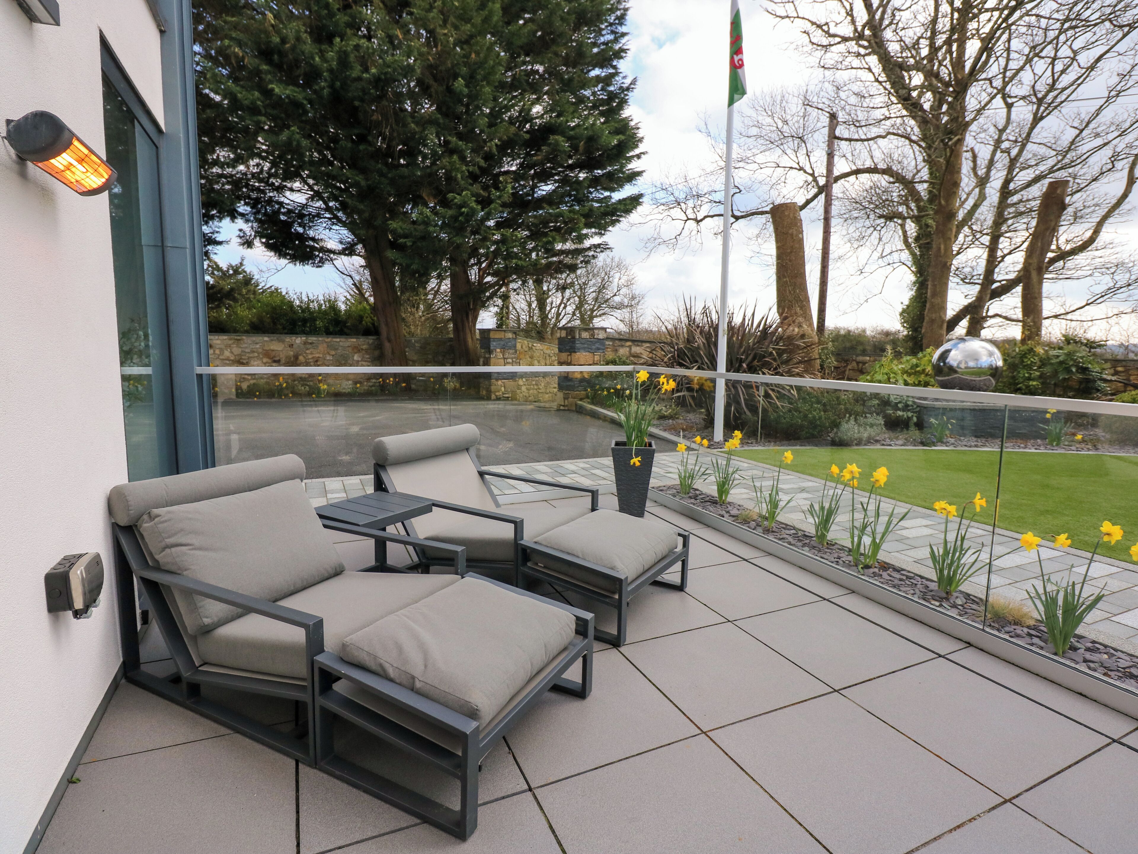 A balcony with two grey cushioned chairs and a small table, a potted plant with yellow flowers, and a clear view of a grassy area with trees and a stone wall in the background.