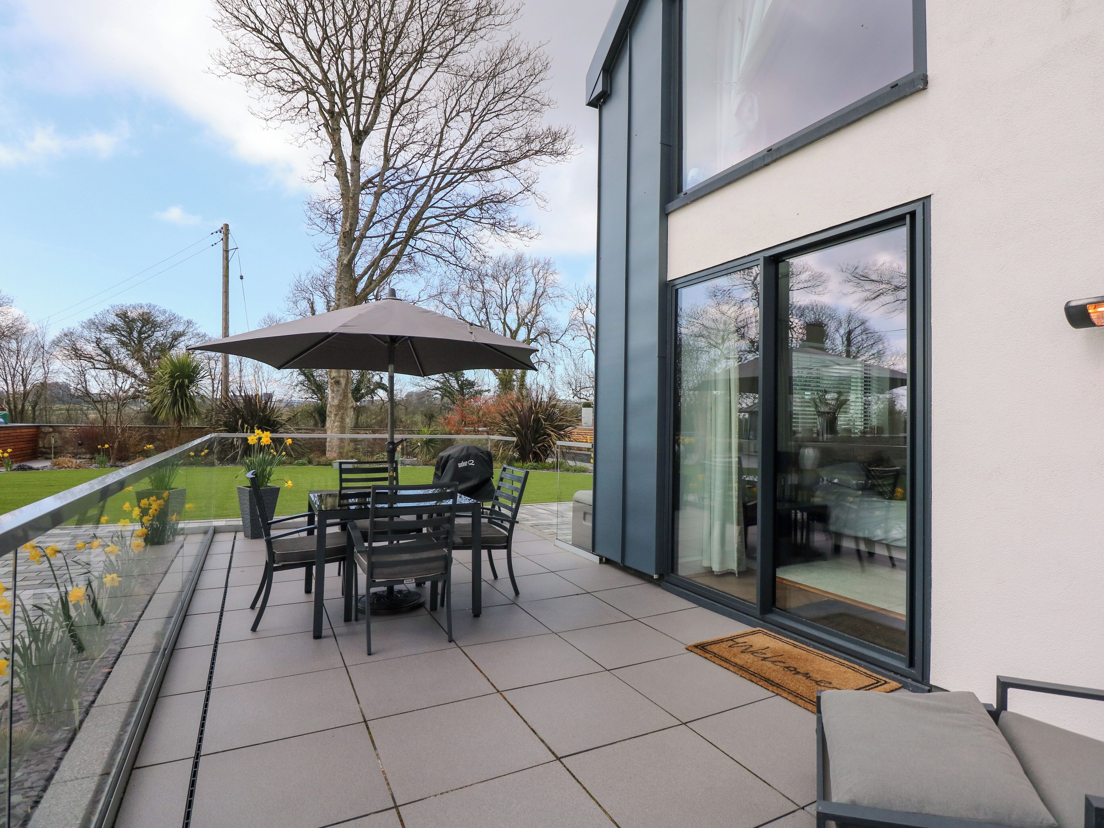Une terrasse extérieure moderne, dotée d’une table et de chaises, d’un grand parasol et d’une porte vitrée donnant sur un jardin.