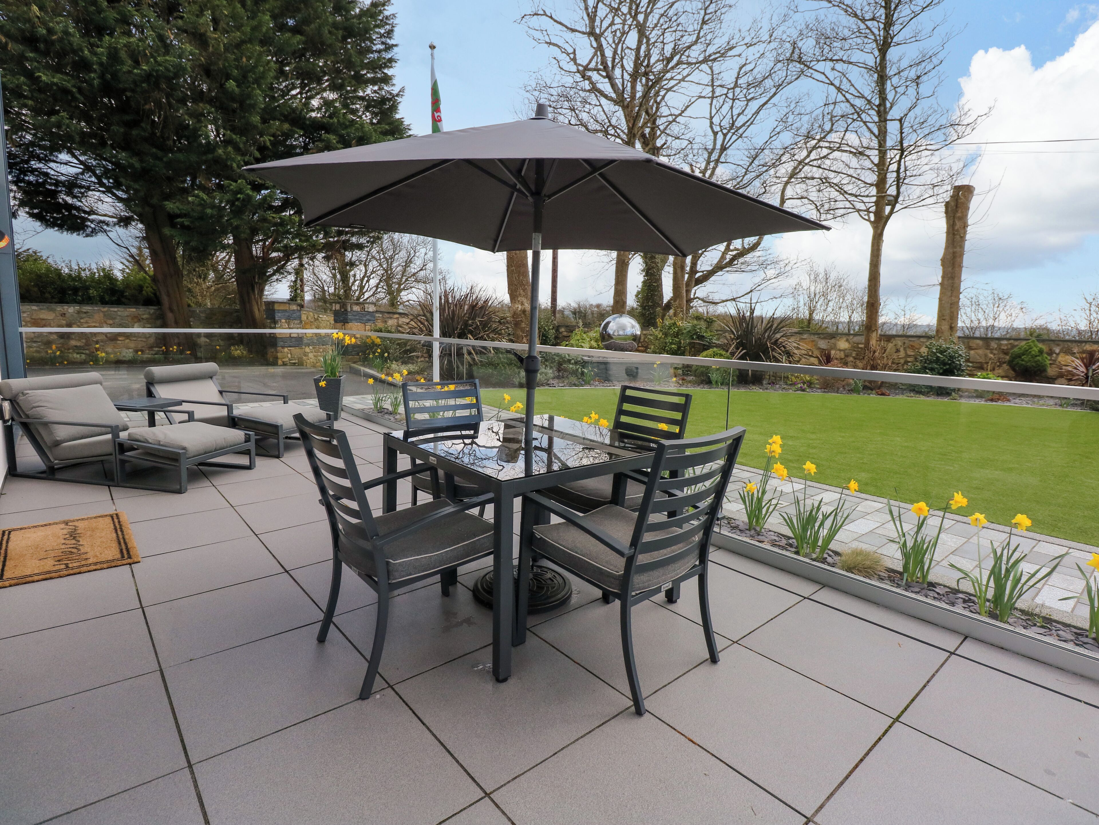 An outdoor patio with a table and chairs, surrounded by a garden with yellow flowers and trees.
