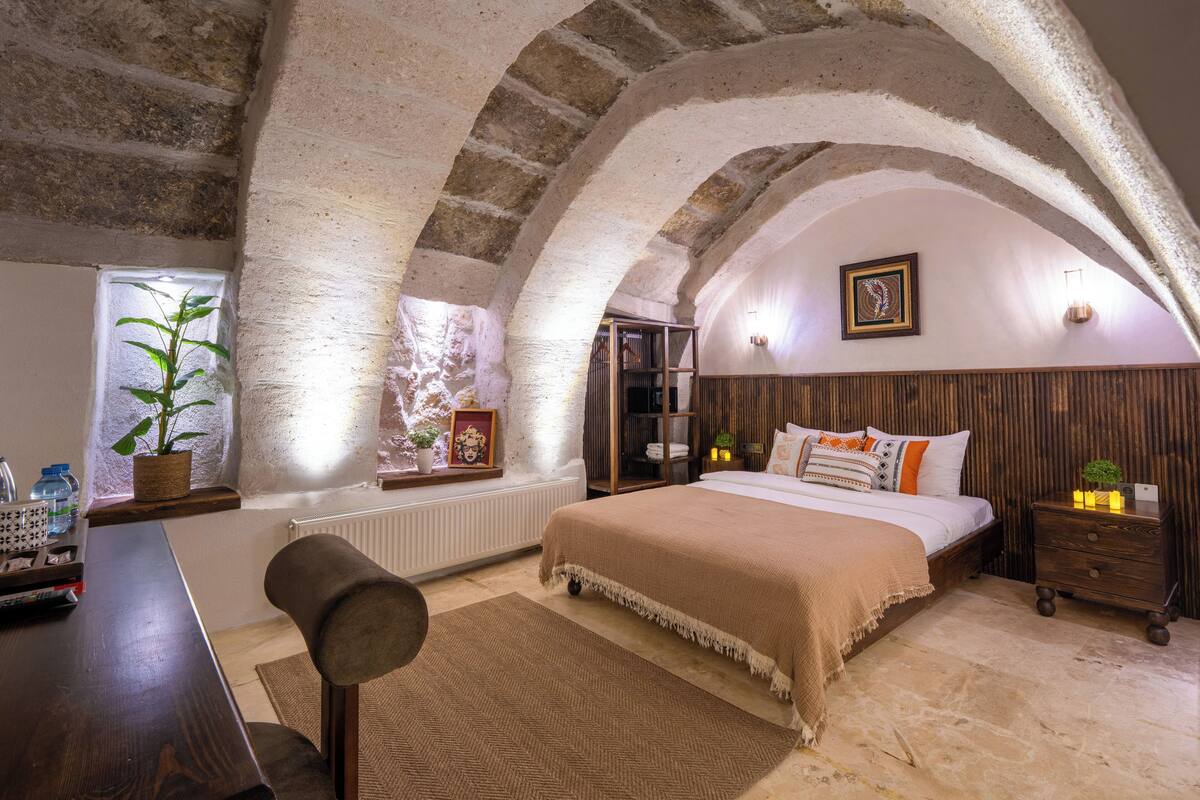 Un dormitorio con techo abovedado, una cama, mesitas de noche, un radiador y una ventana con una planta.