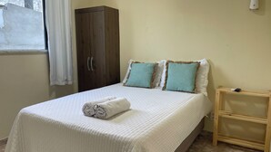 A neatly made bed with a rolled towel, two pillows, a wooden wardrobe, and a small wooden nightstand with a remote control. - Loft CeoPremium 2 | near beaches and shopping malls (Salgado Filho Aracaju)