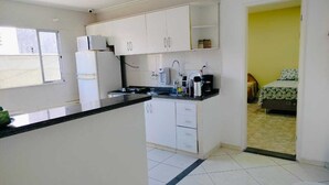 A compact kitchen with white cabinets, a black countertop, and a small sink. A refrigerator and microwave are visible. The room has a tiled floor and a doorway leading to a bedroom with a bed and a yellow wall. - Loft CeoPremium 2 | near beaches and shopping malls (Salgado Filho Aracaju)