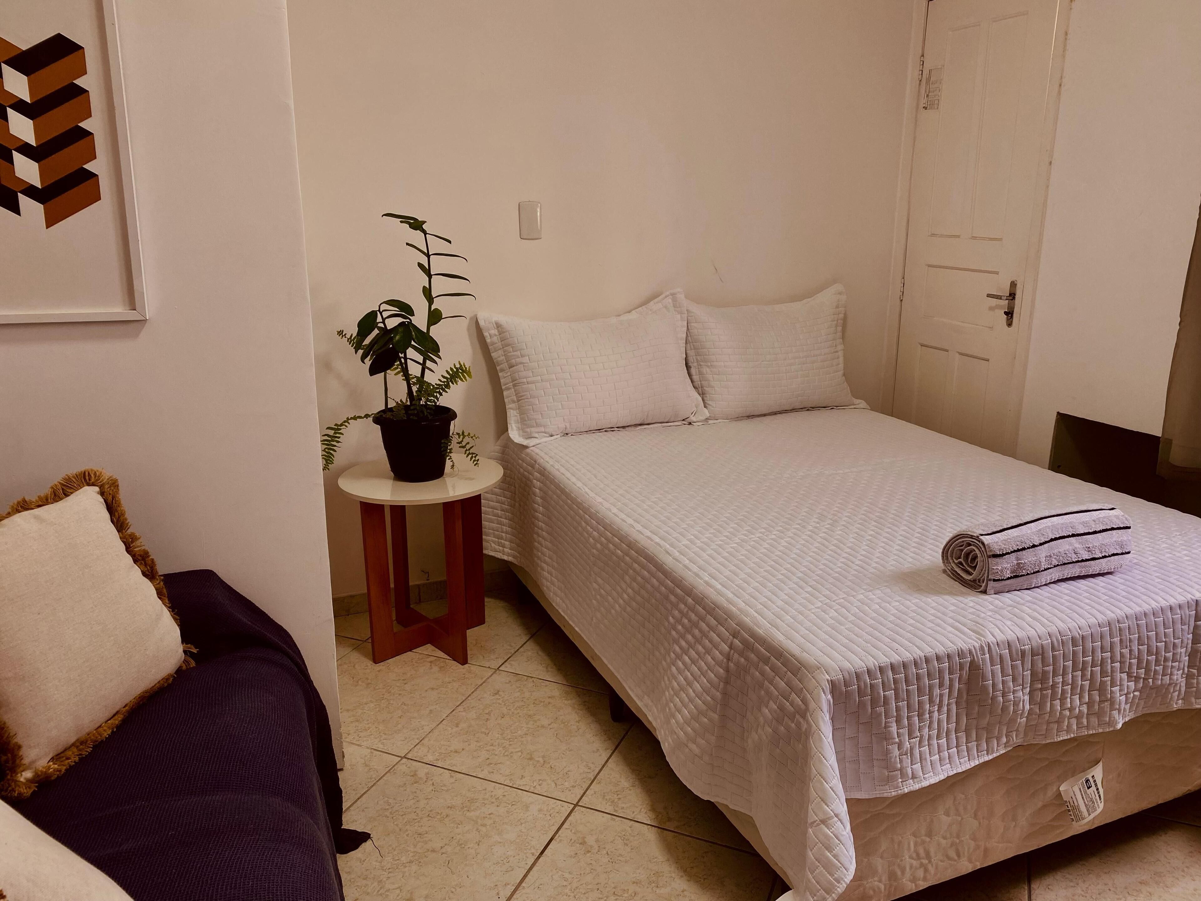 Une chambre bien rangée, avec un lit, une petite table sur laquelle est posée une plante en pot et une chaise.