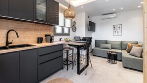 Eine moderne Küche mit einer zentralen Kochinsel, schwarzen Schränken und einer Holz Arbeitsplatte. Es gibt einen Essbereich mit Tisch und Stühlen und ein graues Sofa im Wohnzimmer.