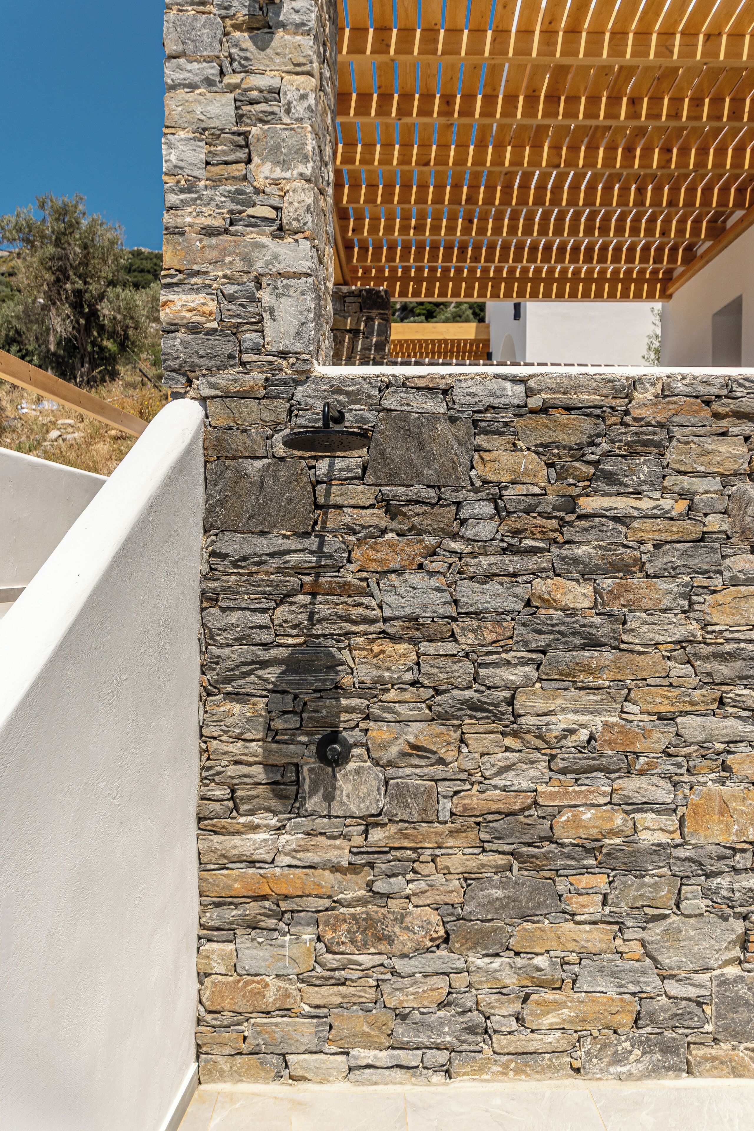 Un muro de piedra con una fuente integrada, una pérgola de madera y una barandilla blanca.