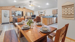 Un comedor amplio con una mesa de madera dispuesta para cuatro, un ventilador de techo y una cocina al fondo.
