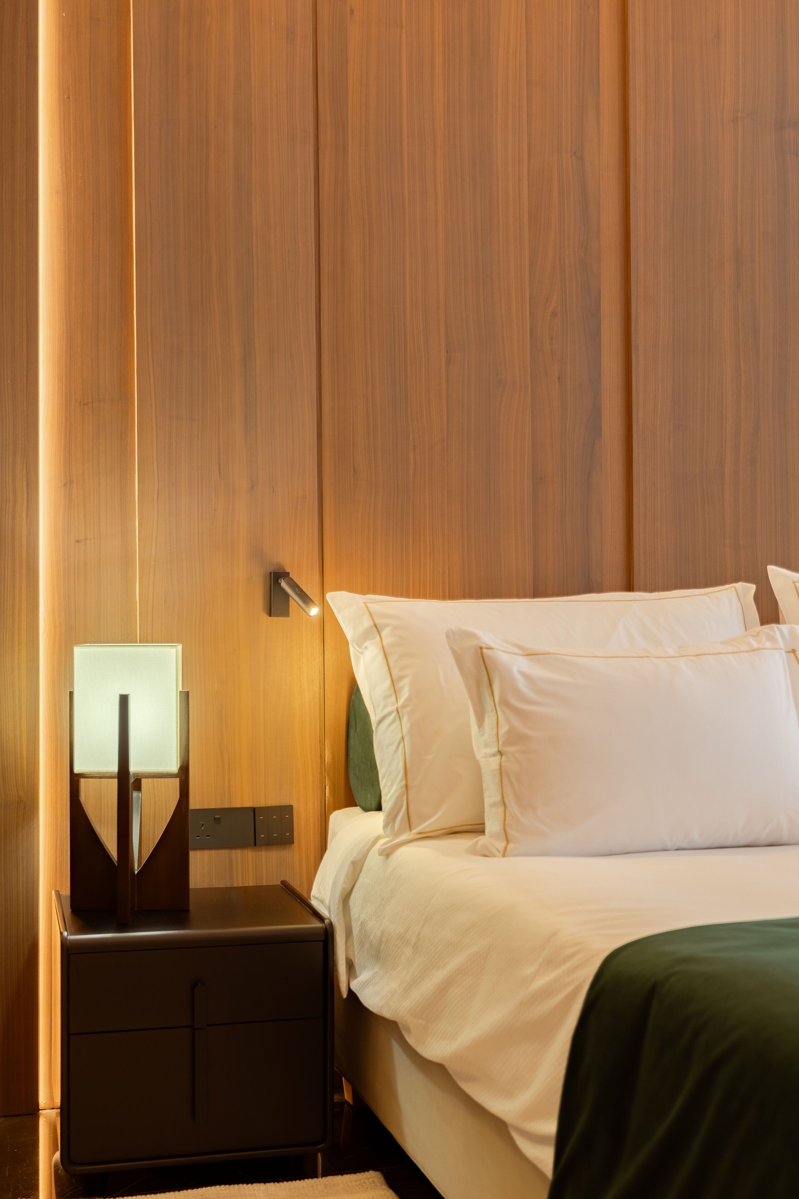 Habitación de hotel con pared de paneles de madera, cama con ropa blanca, mesita de noche con lámpara y manta verde.