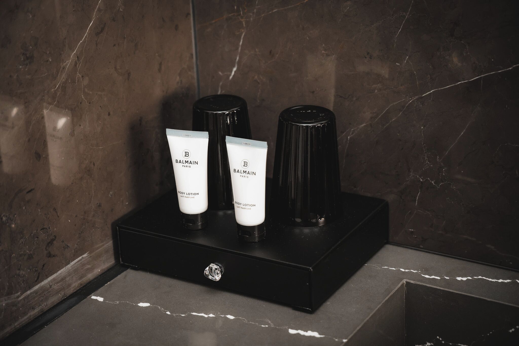 A black tray with three Balmain body lotion tubes and two black cylindrical containers on a marble surface.