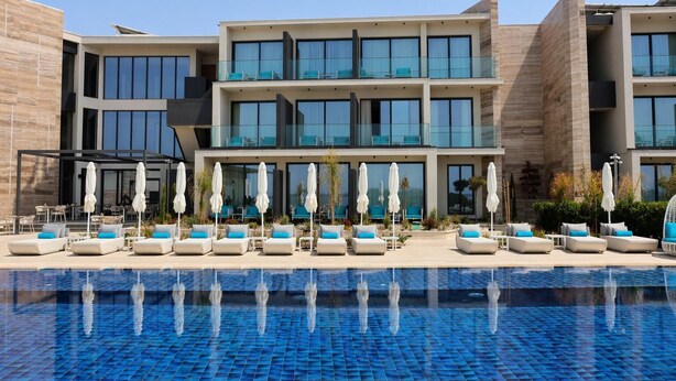 Un hotel moderno con piscina, varias zonas de descanso y un cielo azul despejado.