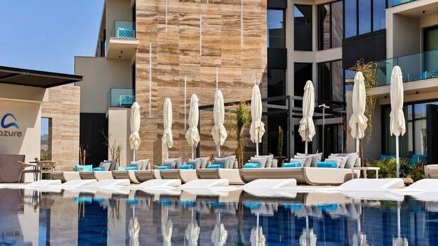Une piscine avec des chaises longues et des parasols, un bâtiment moderne doté d’une façade en pierre et un panneau indiquant le mot « azur ».