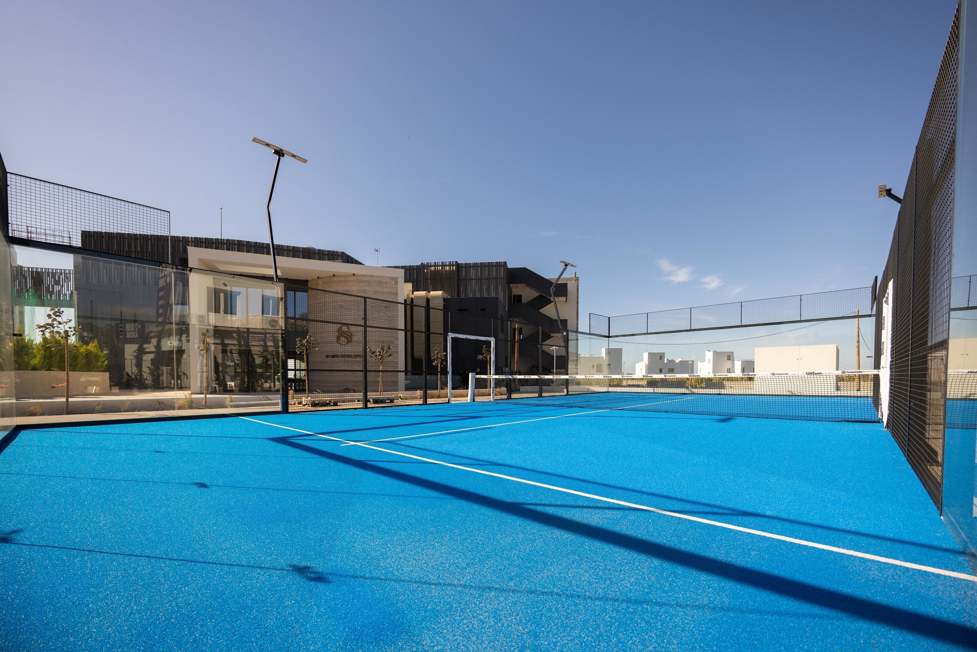 An outdoor tennis court with a net, surrounded by a fence, and adjacent to a modern building with large windows.
