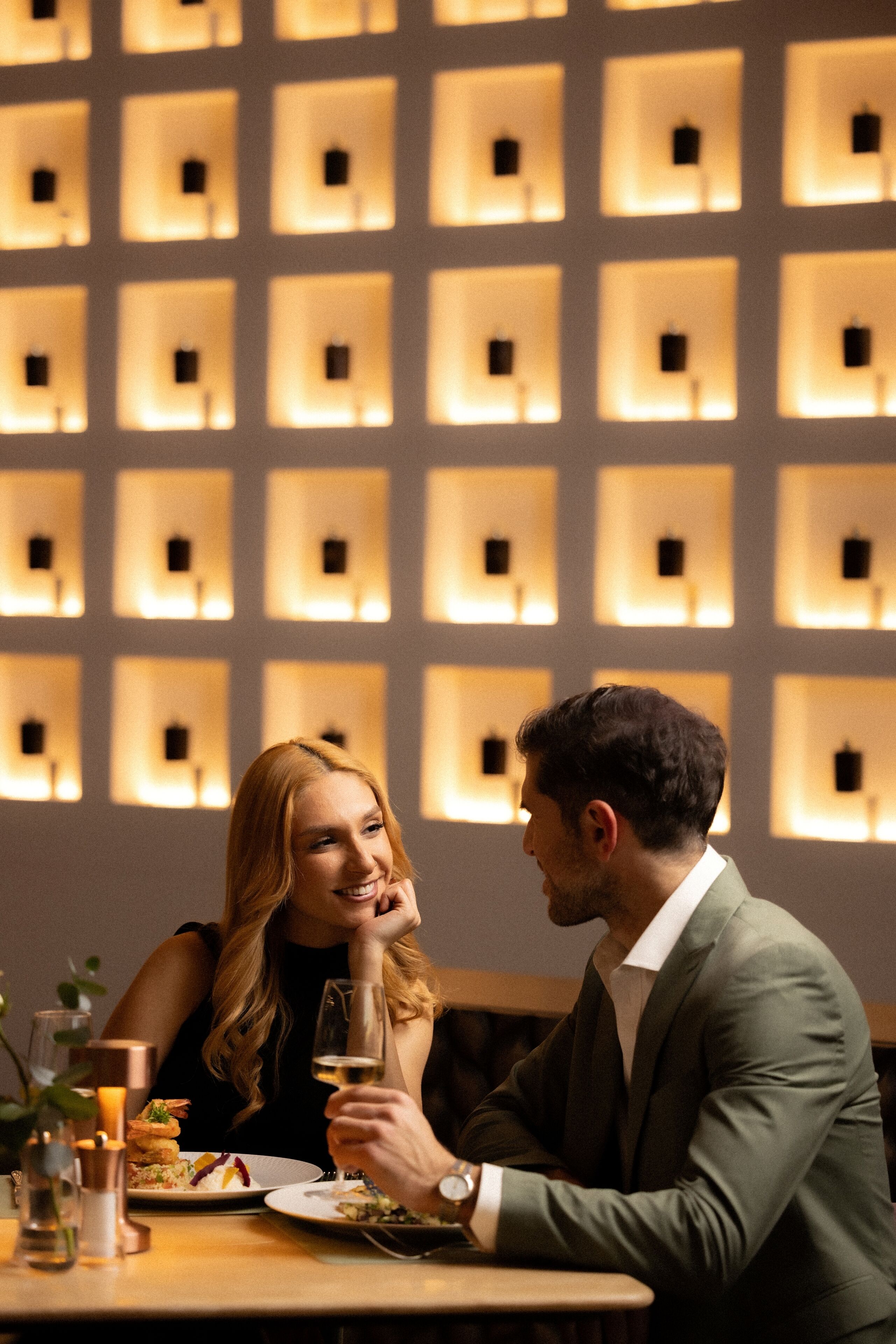 A couple dining at a table with a modern interior featuring recessed lighting and a patterned wall.
