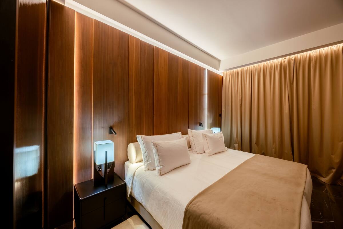 Habitación de hotel con pared de paneles de madera, una cama grande con ropa blanca, una mesita de noche con una lámpara y una cortina en la ventana.
