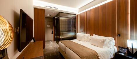 A modern hotel room with a large bed, a flat-screen TV, a desk with a lamp, and a wooden panel wall.