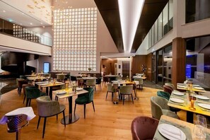 Un restaurante moderno con varias mesas y sillas, un techo alto con iluminación empotrada y una pared grande con diseño de cuadrícula.