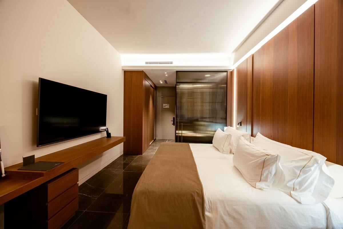 Habitación de hotel moderna con un televisor de pantalla plana grande, un escritorio de madera, una cama con ropa blanca y una puerta corredera que da al baño.